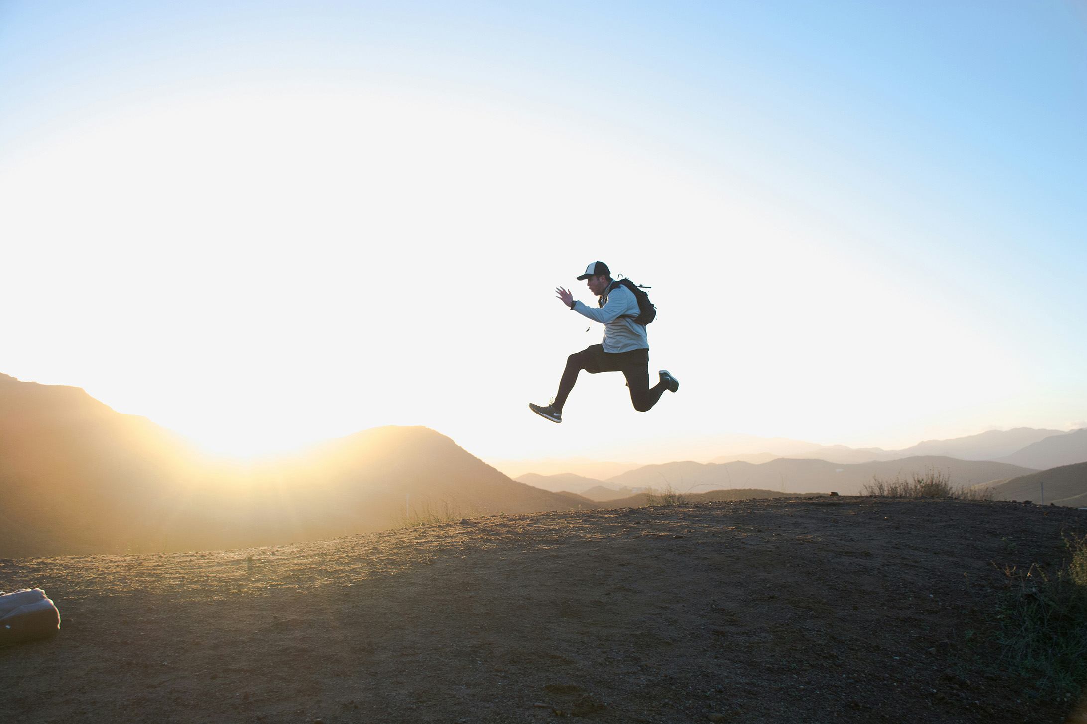 @cam-adams_berghuepfer_unsplash Foto zeigt energiegeladenen Mann bei einem hohen Hüpfer auf einer Hügelkuppe