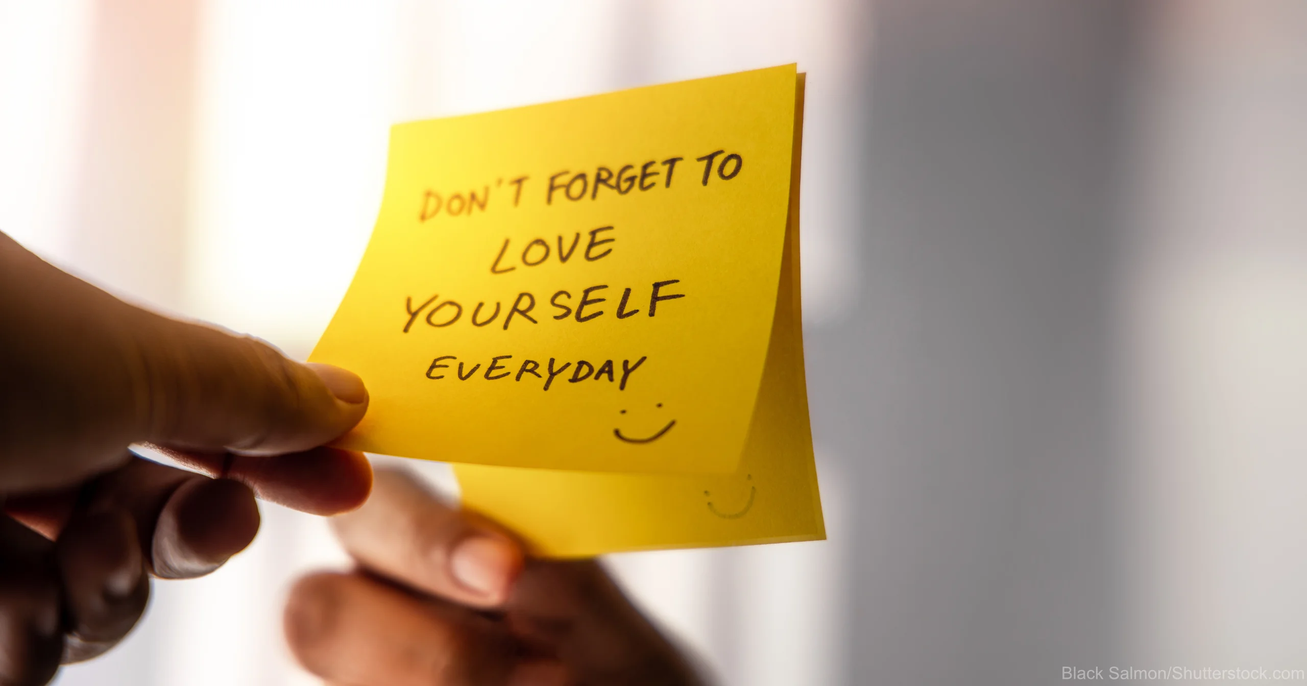 Eine Hand hält einen gelben Notizzettel mit der handschriftlichen Botschaft ‘Don’t forget to love yourself everyday’ und einem kleinen Smiley. Im Hintergrund spiegelt sich die Hand in einem Fenster oder Spiegel, was die Bedeutung von Selbstliebe und Achtsamkeit symbolisiert.